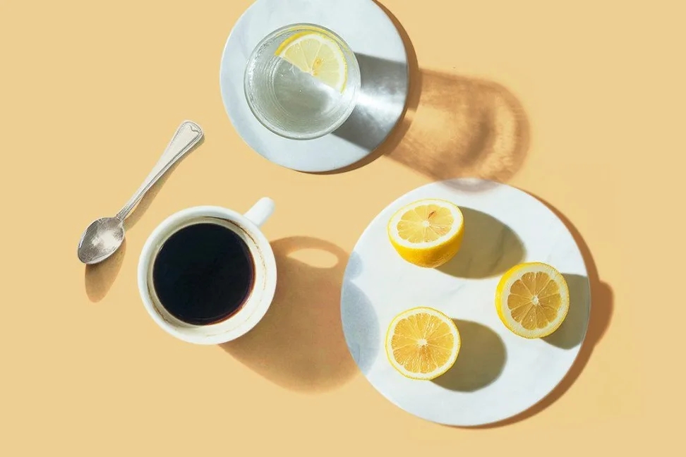 a white plate topped with slices of lemon next to a cup of coffee and a spoon on a yellow background
