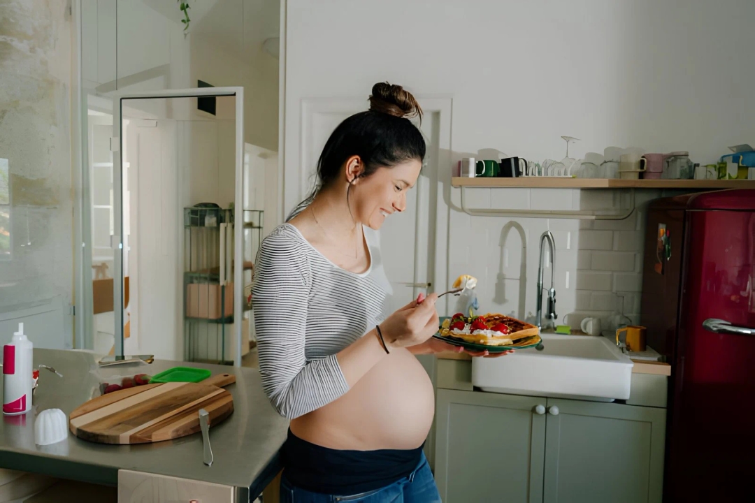 Portrait of a smiling future mother eating waffles in the kitchen of her apartment; craving sweet food during pregnancy.