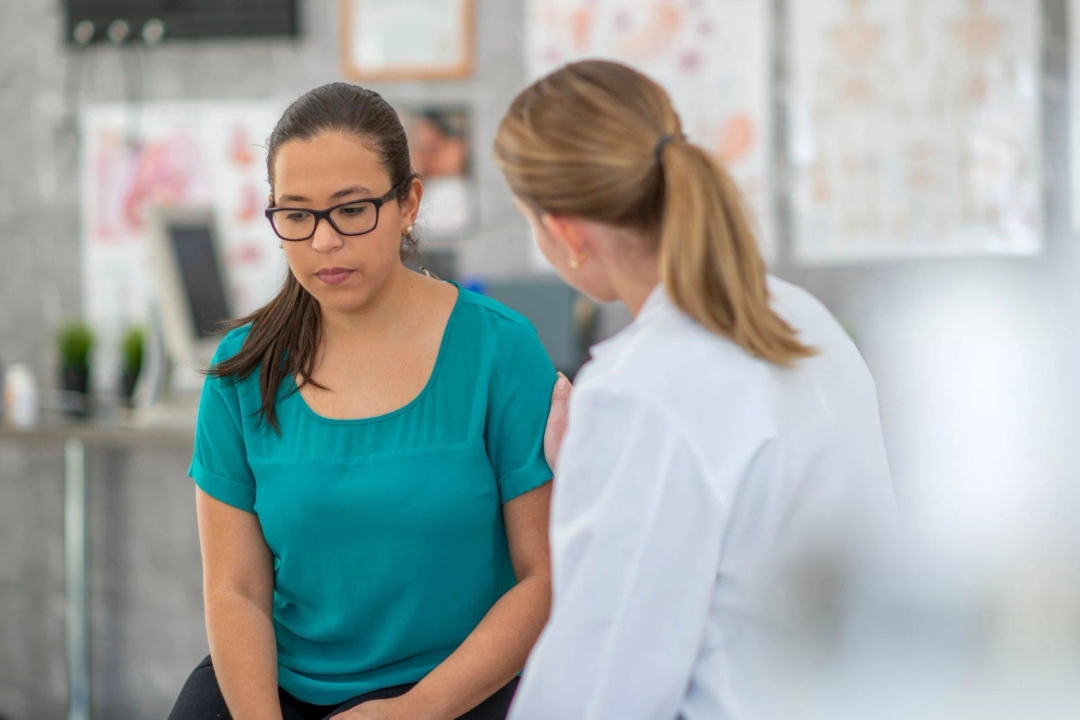 A young woman looks at the floor nervously while her hands rest on her lap. Her doctor sits across from her telling her some bad news