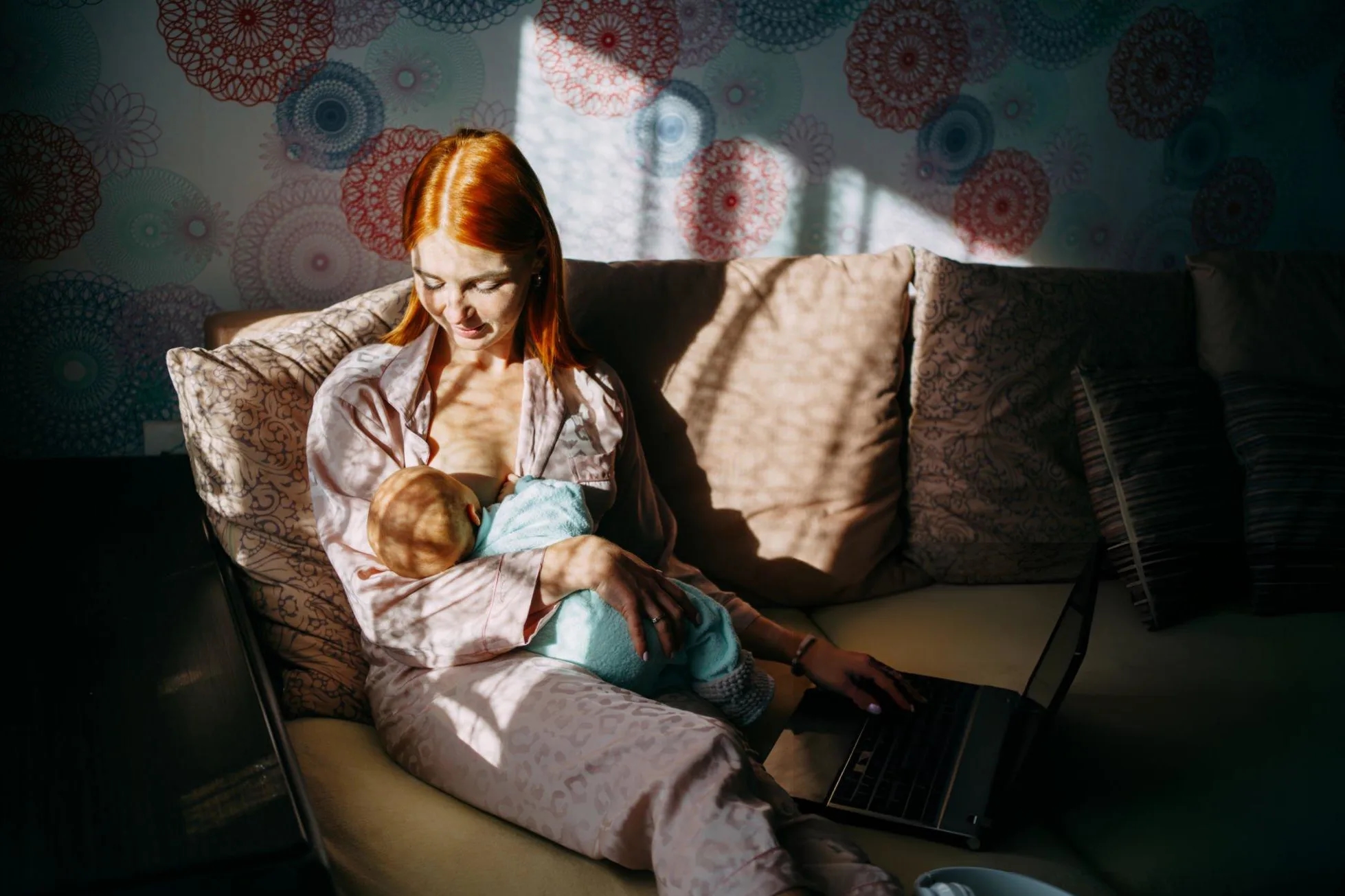 Mother breastfeeds her newborn son and works remotely with a laptop at home.