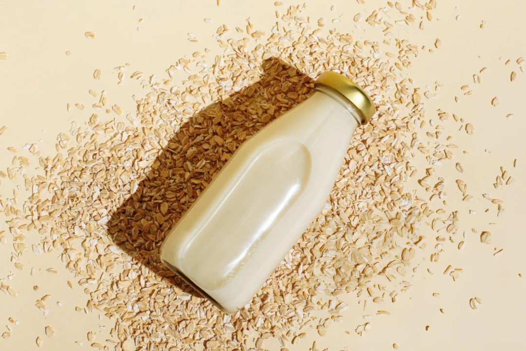 Healthiest Oat Milks - Oat milk in glass bottle and oatmeal flakes on beige background. Healthy vegan non-dairy organic drink with flakes. Lactose-free milks in minimal flat lay style. Top view