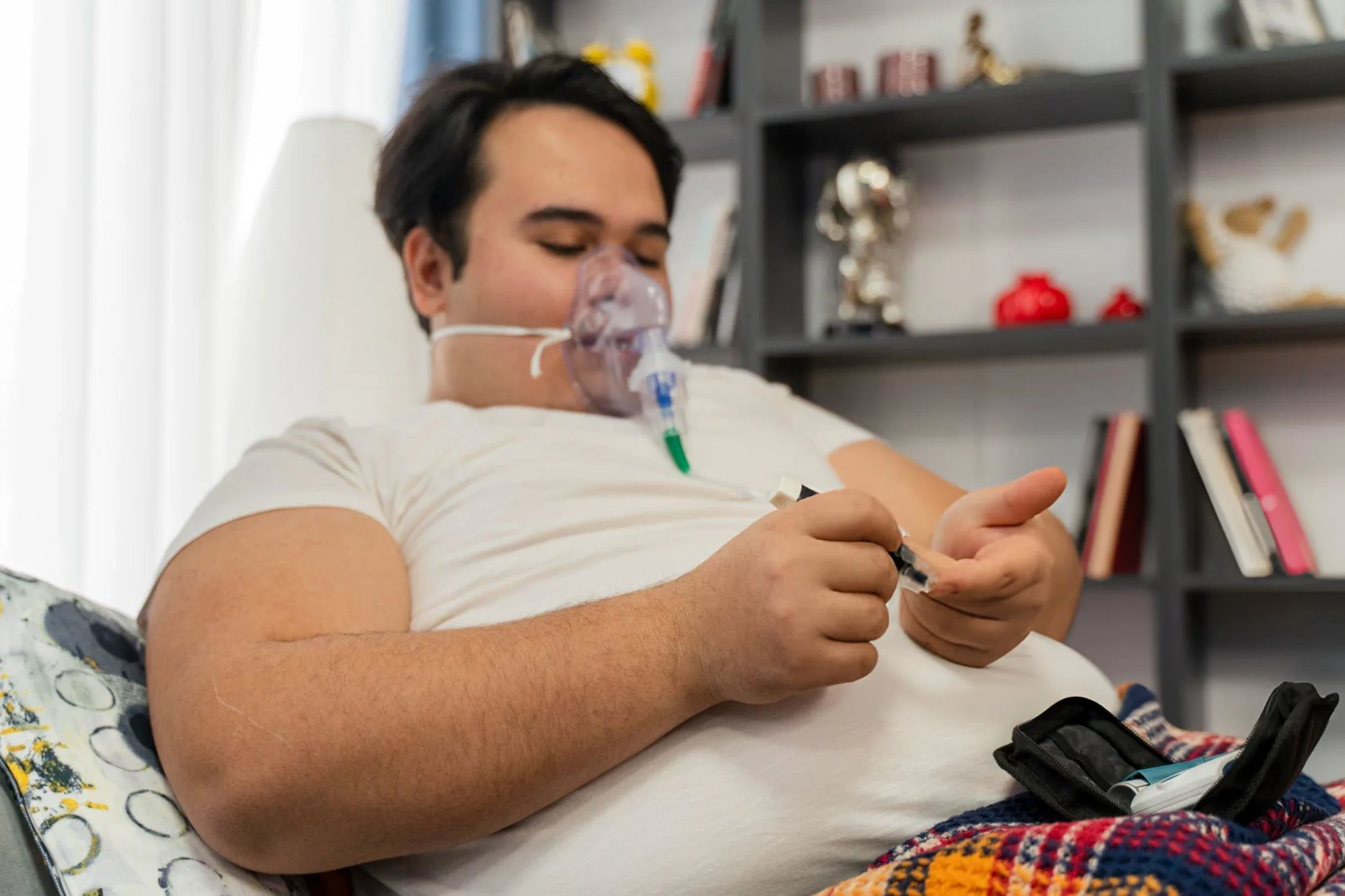 What are the Causes of adult obesity ? Young man sits in a sofa with an oxygen mask and measuring blood sugar level